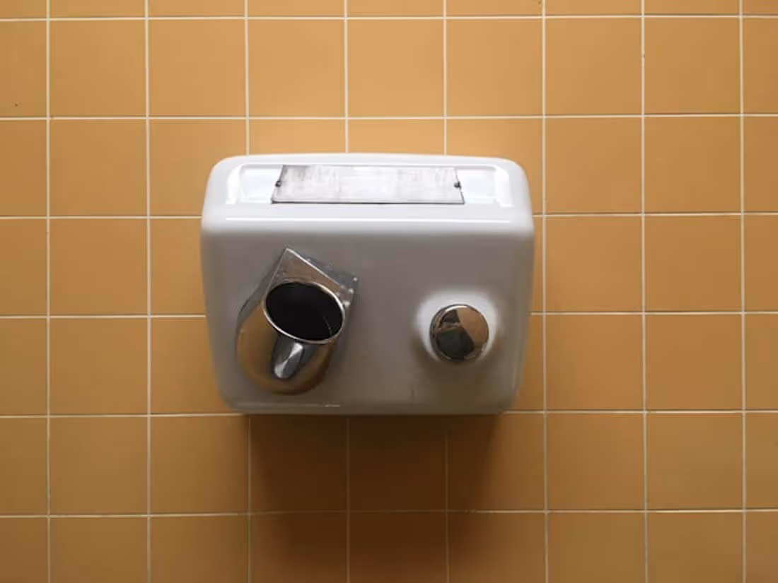 Wall-mounted white hand dryer with a metallic nozzle and button, set against a tan tiled wall.