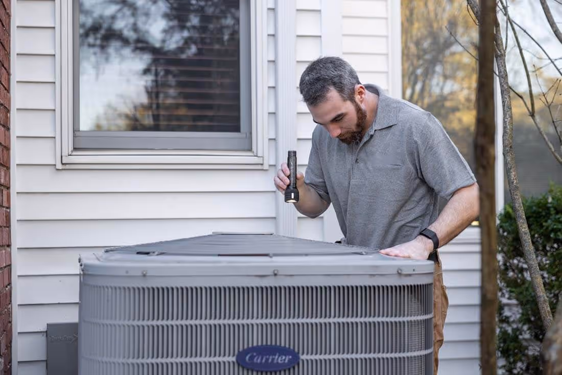 Technician inspecting an outdoor Carrier HVAC unit with a flashlight next to a house.