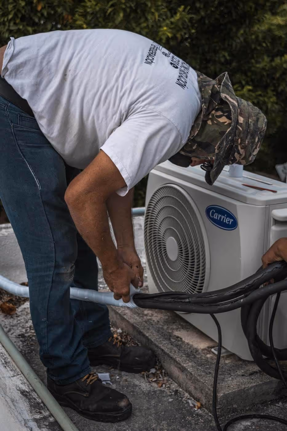 Technician in a camouflage hat and white shirt installing or repairing a Carrier air conditioning unit outdoors.