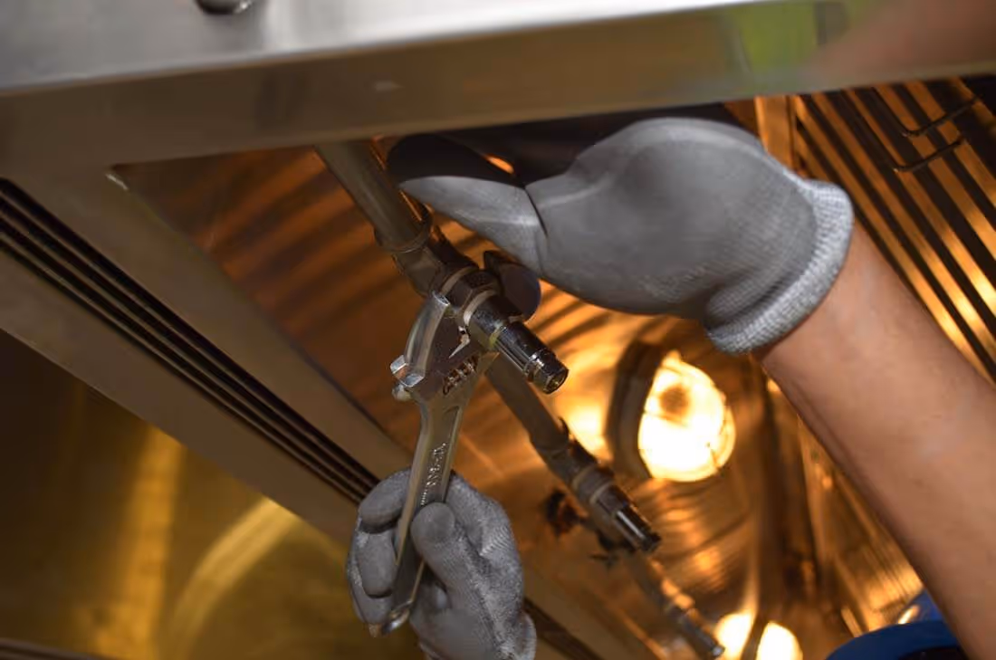 Hands wearing gray gloves using a wrench to tighten a pipe valve under a metal surface with lights in the background.