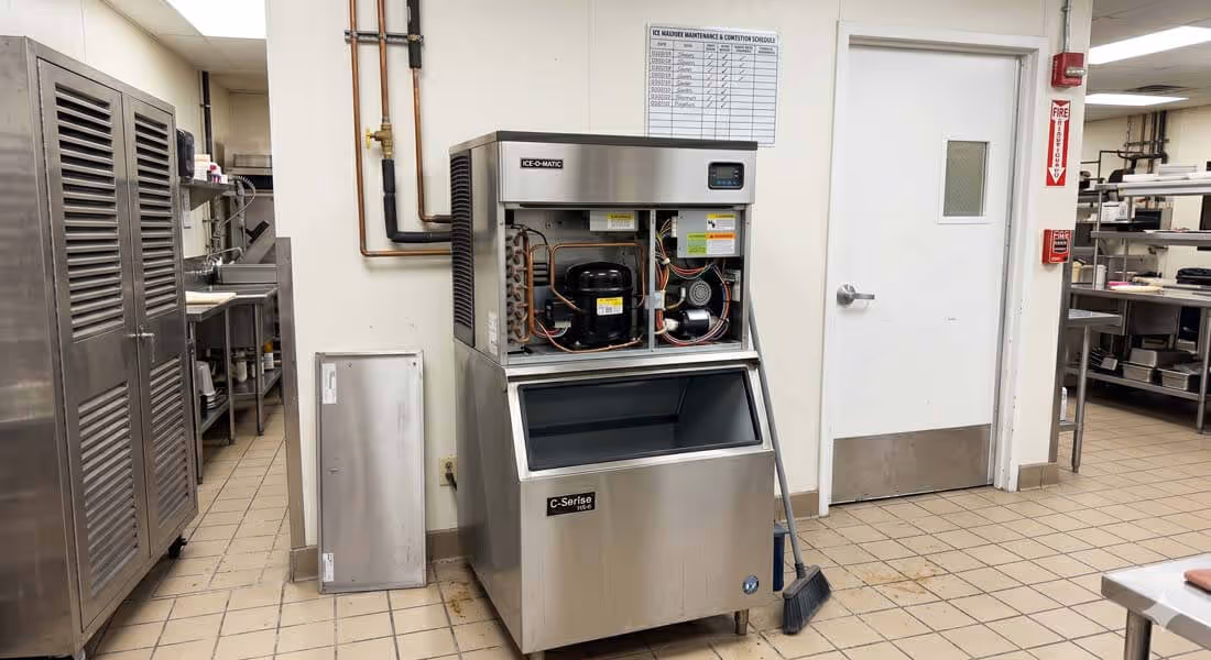 Commercial ice machine with open maintenance panel and broom leaning against it in a kitchen with tiled floor and stainless steel cabinets.