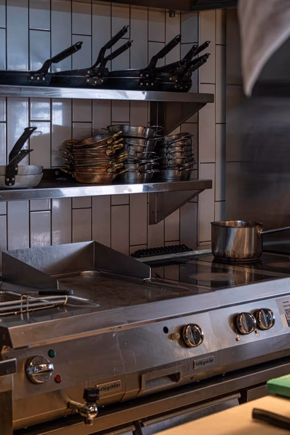 Commercial kitchen stove with knobs and a pot, shelves above holding stacked pans and skillets against white tiled wall.