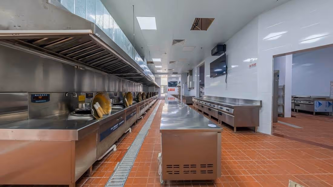 Spacious industrial kitchen with stainless steel counters, cooking stations, and orange tiled floor.